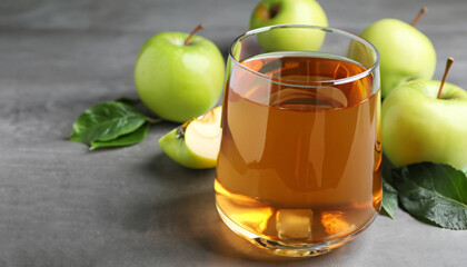 Glass of delicious cider, green leaves and apples on gray table, closeup. Space for text