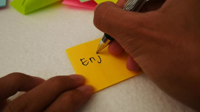 Enjoy every moment- Man writing "Enjoy every moment" on a pink notepaper with a black pen.