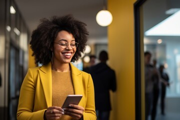 A woman stands proudly indoors, wearing her favorite outfit and smiling brightly as she holds her phone, her joy radiating from the wall behind her