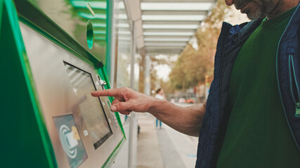 Mature man dressed in casual clothes buys ticket in the electronic terminal while standing at public transport stop