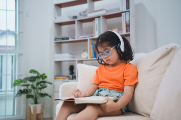 Asian baby girl child wearing glasses and headphone write notes in notebook and reading book study...