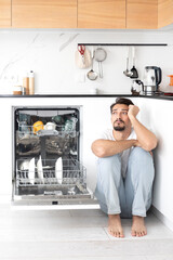 Tired and bored man putting dirty plates in dishwasher machine in the kitchen. Household and...