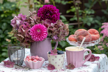 Tisch-Arrangement mit frischen Heidelbeeren und Pfirsichen, Tasse Kaffee, Süßigkeiten und Blumenstrauß mit Dahlien und Hortensienblüten
