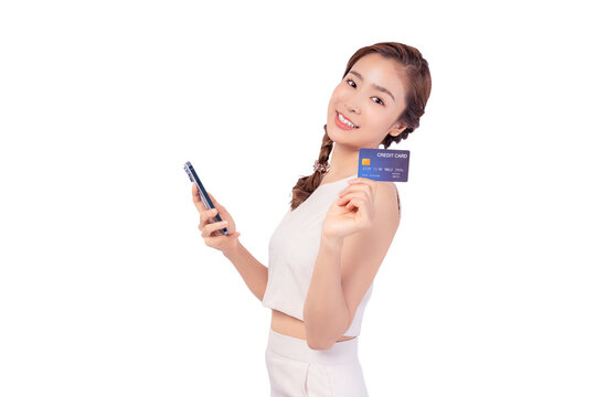 Young asian lady using mobile phone holding credit card looking at camera isolated over pastel white background and copy space Smiling young girl purchasing online through phone using credit card