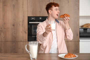 Fototapeta premium Young redhead man with glass of milk eating croissant in kitchen