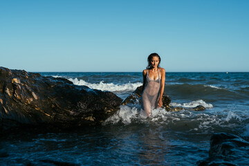 A beautiful wet brunette girl in a silvery net stands in the water on the seashore