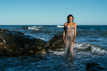 Fototapeta na wymiar A beautiful wet brunette girl in a silvery net stands in the water on the seashore