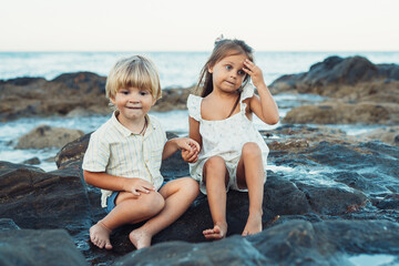 Fototapeta na wymiar blond boy and a brunette girl, three years old, are sitting on stones on the seashore