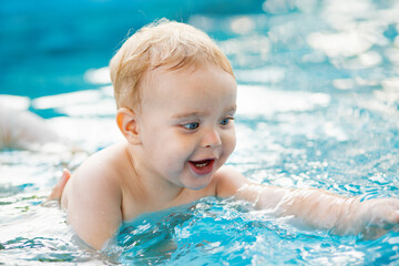 Portrait of small red-haired boy bathes in pool with hand support, baby swimming in water, summer leisure