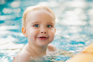 Fototapeta na wymiar Portrait of small red-haired boy bathes in pool with hand support, baby swimming in water, summer leisure