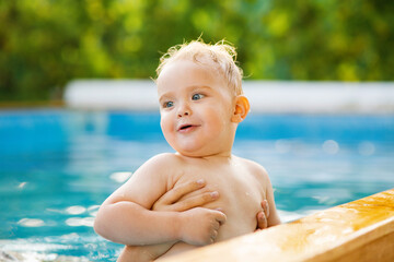 Fototapeta na wymiar Portrait of small red-haired boy bathes in pool with hand support, baby swimming in water, summer leisure