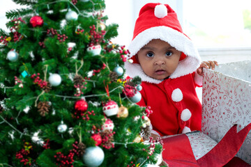Cute baby African girl in red costume standing in big box near Christmas tree, looking at camera