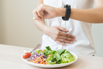 Intermittent fasting, health care asian young woman dietary, having stomach ache, temptation hungry of breakfast food in morning on table at home, looking at watch on her wrist but not yet time to eat