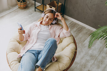 Top view young woman wear casual clothes sits in archair listen to music in headphones using mobile cell phone stay at home hotel flat rest spend free spare time in living room indoor. Lounge concept.