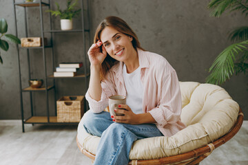 Obraz premium Smiling happy cheerful fun young woman wear casual clothes sits in armchair drink coffee tea stay at home hotel flat rest relax spend free spare time in living room indoor. Lifestyle lounge concept.