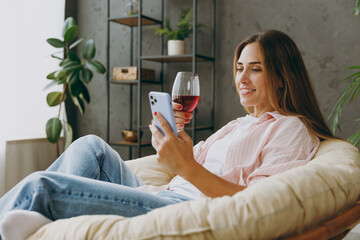 Side view young woman wear casual clothes sits in armchair hold wineglass drink wine use mobile cell phone stay home hotel flat rest relax spend free spare time in living room indoor Lounge concept. - Powered by Adobe