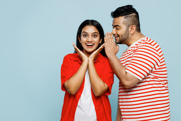 Young couple two friends family Indian man woman wear red casual clothes t-shirts together whispering gossip and tells secret behind his hand sharing news isolated on plain blue cyan color background.