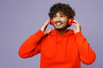 Young fun Indian man wears red orange hoody casual clothes listen to music in headphones look aside...