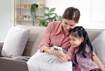 Millennial Asian happy family mother sitting smiling on cozy sofa couch holding showing funny content streaming online via touchscreen smartphone to little young girl daughter in living room at home.