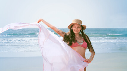 young woman in a bikini stands by the sea with a flowing towel