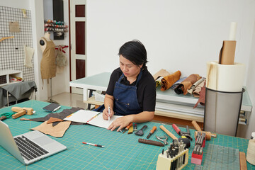 Asian Female Leather worker using pen to make sketch on notebook at the studio 