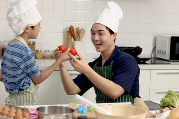 Happy Asian father and son kid with apron and chef hat holding red tomato during cooking healthy...