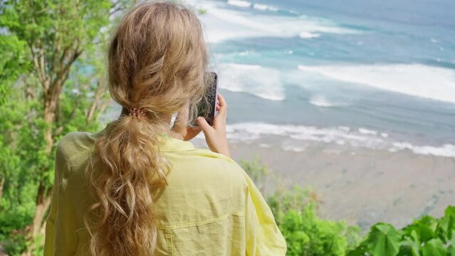 A woman tourist in yellow shirt shoots a video on the phone, a Back view, a Blonde girl photographs the ocean and nature of Bali, Green bowl beach. Slow motion 