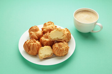 Homemade profiteroles with cream on plate with cup of coffee on mint background. Fresh baked golden profiterole.