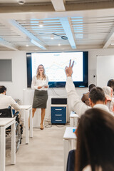 Senior businessman raising a hand to ask a question while his blonde, female, business trainer explaining something