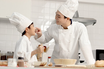 Happy Asian son and father in chef uniform with hat at kitchen, parent and boy child prepare bread dough, dad touches flour on son nose by finger, cute family have fun together during making bakery.