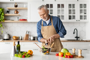 Happy senior man preparing healthy vegetable salad in kitchen, seasoning meal