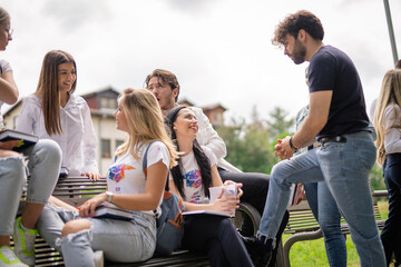 Young, happy, college students having a conversation sitting on a bench in the park out of the campus building, discussing school subjects or project, preparing for exam