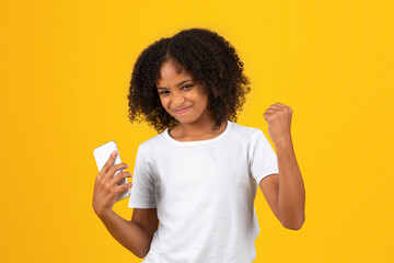 Cheerful adolescent african american girl in white t-shirt, uses phone celebrate success
