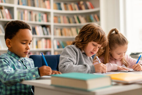 Smart diverse schoolchildren sitting at table in classroom at primary school, writing or drawing in notebooks
