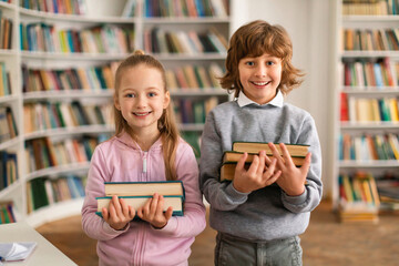 Portrait of happy european boy and girl pupils holding a stack of books, standing in library and...