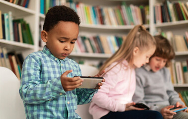 School children playing video games on smartphones during break while sitting in library or classroom, selective focus