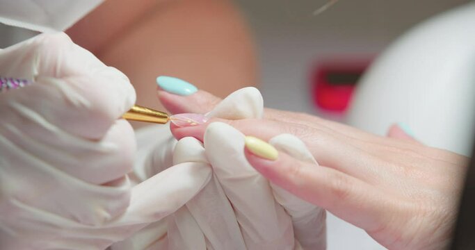 Closeup view of female caucasian hands. The manicurist in gloves draws yellow ornament on the client's nails. Elegant nails design. Cosmetology and beauty studio.