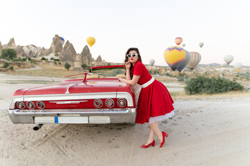 beautiful girl in retro style posing near a vintage red cabriolet car on background of balloons in Cappadocia. - obrazy, fototapety, plakaty