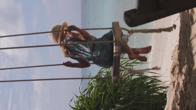 Vertical video. Little girl in blue sundress and straw hat swinging on a beach swing over scenic view with turquoise ocean water, slow motion