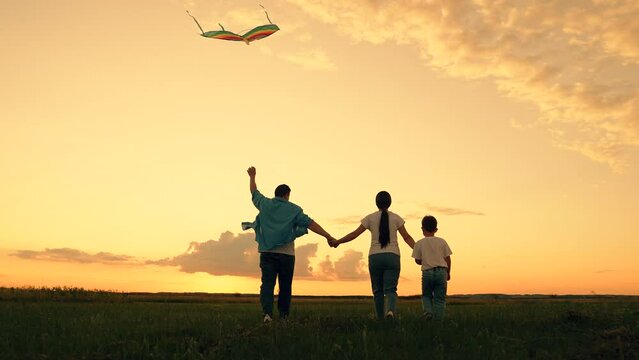 Young family is playing with kite in field. Happy family with baby in park playing with kite. Family walk on grass, child dreams of flying. People together travel, nature, silhouette. Kid wants to fly