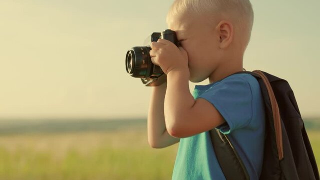 Boy with backpack on back look into camera lens, take beautiful photos of field with flowers. Kid with camera in hands take photo nature on sunny day. Young photographer in fresh air film about nature