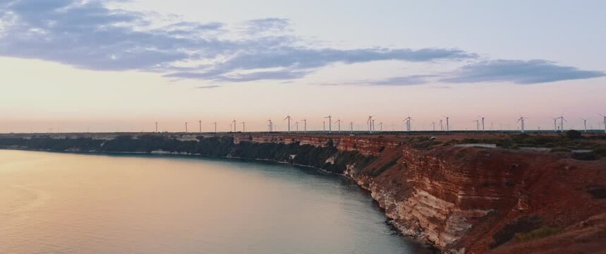 cinematic flight over a cape on the seashore at sunset, on the horizon many wind turbines generate green energy. Anamorphic 2.39 scope