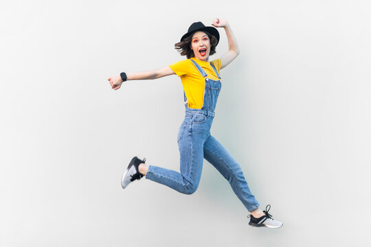 Portrait of excited positive hipster woman in blue denim overalls, yellow T-shirt and black hat, jumping in the air, moving, expressing happiness. Indoor studio shot isolated on gray background.