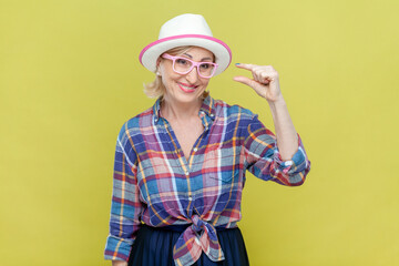 Portrait of senior woman wearing checkered shirt, hat and eyeglasses shows something very small or tiny, discusses prices in store, smiles happily. Indoor studio shot isolated on yellow background.