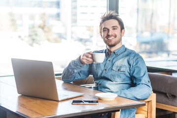 Fototapeta na wymiar Portrait of delighted smiling happy young man freelancer in blue jeans shirt working on laptop, having break, drinking coffee. Indoor shot near big window, cafe background.