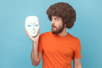 Portrait of man with Afro hairstyle wearing orange T-shirt looking at white mask in hands with...