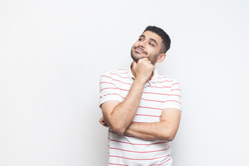 Portrait of pensive dreaming positive bearded man wearing striped t-shirt standing looking away, thinking about something pleasant. Indoor studio shot isolated on gray background.