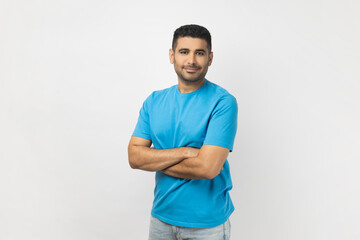 Fototapeta premium Portrait of handsome joyful unshaven man wearing blue T- shirt standing looking at camera, expressing positive emotions, keeps arms crossed. Indoor studio shot isolated on gray background.
