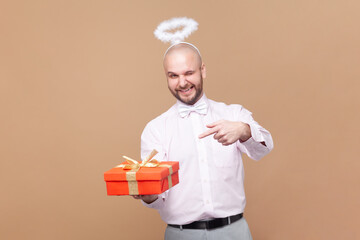 Cute optimistic bald bearded man with nimb over head, holding pointing at red present box, looking...