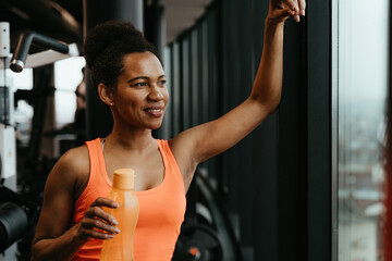 Fit African American woman standing next to the window and holding water bottle while smiling and...
